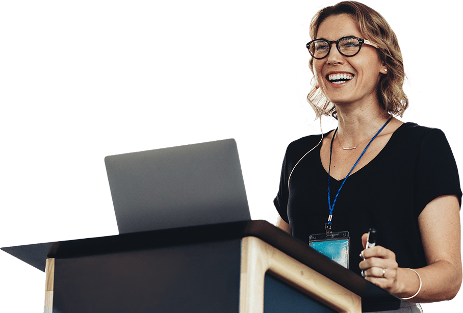 Person speaking at a podium with a laptop and microphone, smiling and wearing glasses.
