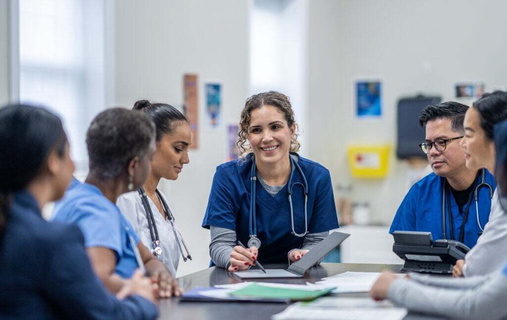 A group of healthcare professionals in scrubs sit around a table in an academic clinic setting, having a discussion.