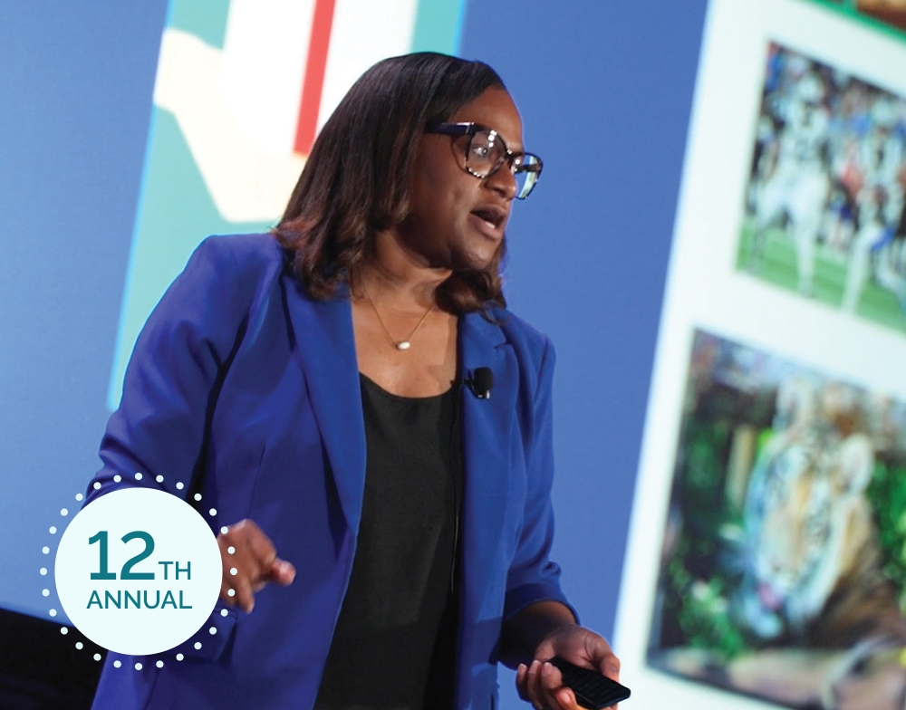 A woman in a blue blazer speaks on stage at the PSIvet Business Symposium. A "12th Annual" badge is visible.